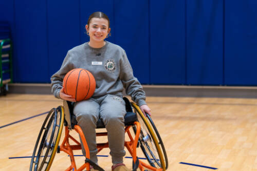 student in a wheelchair holds a basketball on the court and smiles at the camera